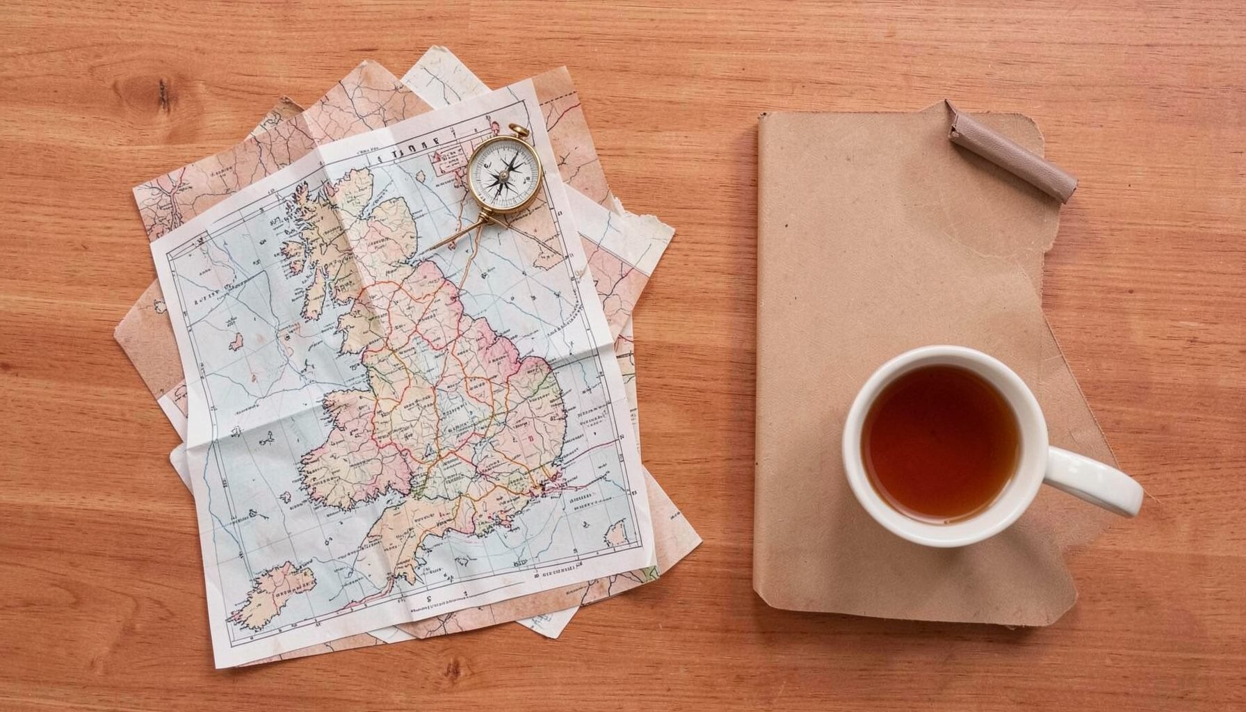 Overhead flat-lay photograph of travel essentials on a warm wooden surface: a folded map of Ireland, a vintage compass, and a notebook